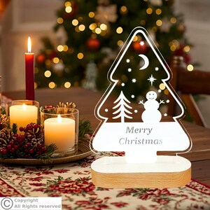May include: A clear acrylic Christmas tree-shaped ornament with a white base. The design features a snowman, a small tree, a crescent moon, and stars, with the text Merry Christmas printed in the center of the tree shape.