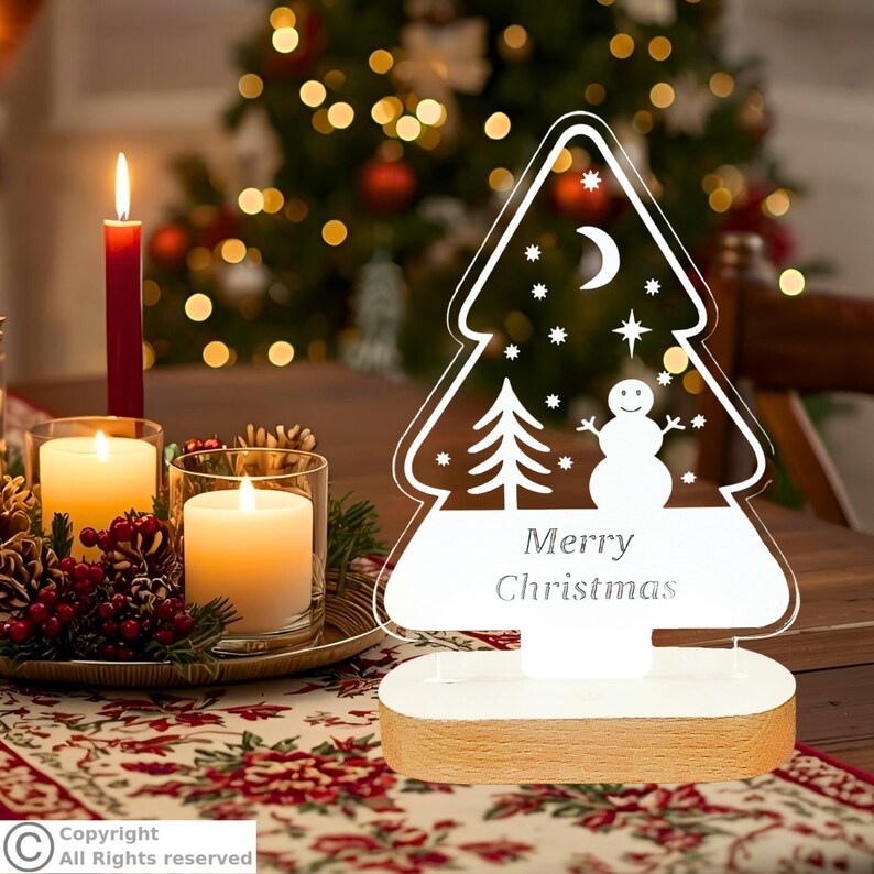 May include: A clear acrylic Christmas tree-shaped ornament with a white base. The design features a snowman, a small tree, a crescent moon, and stars, with the text Merry Christmas printed in the center of the tree shape.
