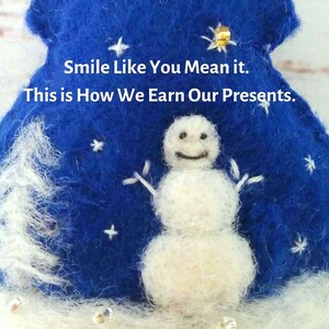 May include: A blue felted ornament featuring a white needle-felted snowman, embroidered white stars, and a gold sequin. The text reads: Smile Like You Mean it. This is How We Earn Our Presents. The base is white with small clear beads.