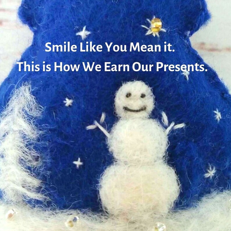 May include: A blue felted ornament featuring a white needle-felted snowman, embroidered white stars, and a gold sequin. The text reads: Smile Like You Mean it. This is How We Earn Our Presents. The base is white with small clear beads.