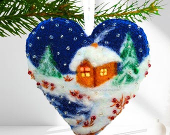 May include: A heart-shaped felt ornament featuring a dark blue night sky with scattered clear beads, a small orange house with glowing yellow windows covered in white snow, green pine trees, and red berries, suspended by a white ribbon.