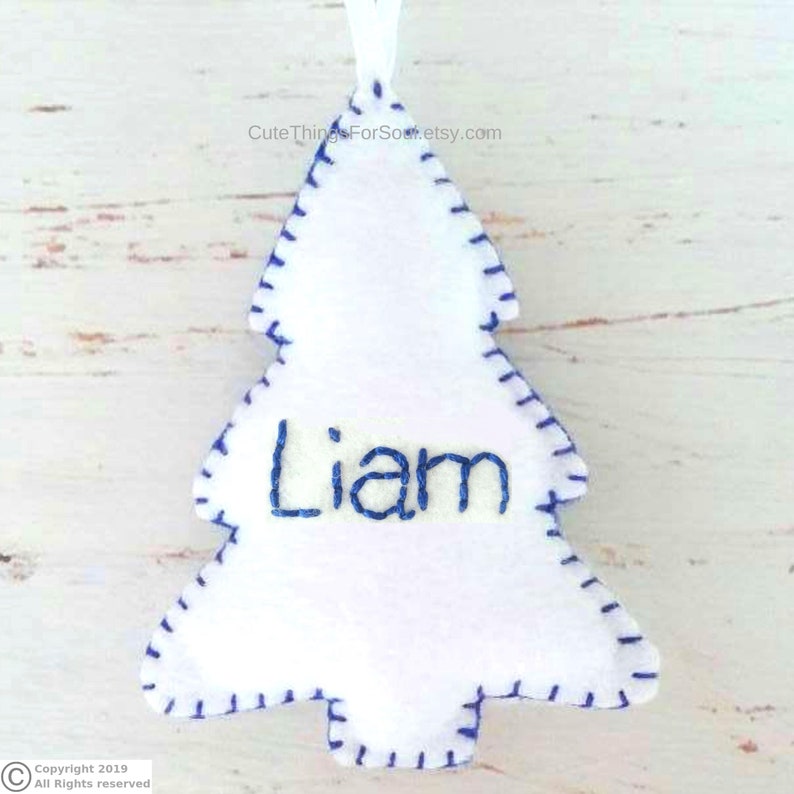 May include: A white felt Christmas tree ornament featuring a blue stitched border and the name Liam embroidered in blue thread across the center. A small white ribbon loop is attached to the top for hanging purposes.