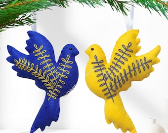 May include: Two felt bird ornaments hanging from white ribbons. One bird is blue with yellow embroidered leaf patterns on its wing, and the other is yellow with blue embroidered leaf patterns on its wing. Both birds have small black bead eyes.