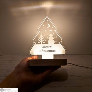 May include: A clear acrylic Christmas tree-shaped night light with a wooden base. The design features an etched snowman, a small tree, stars, and a crescent moon. The text Merry Christmas is centered on the lower portion of the tree.