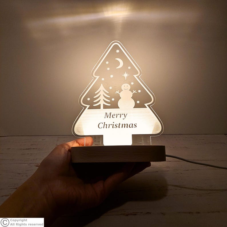 May include: A clear acrylic Christmas tree-shaped night light with a wooden base. The design features an etched snowman, a small tree, stars, and a crescent moon. The text Merry Christmas is centered on the lower portion of the tree.