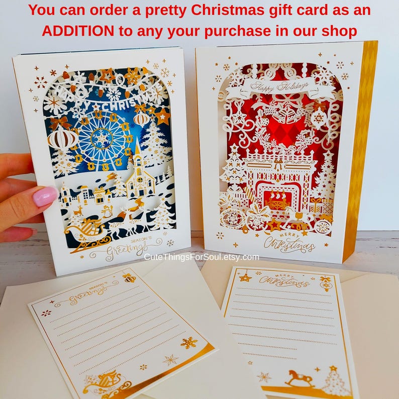May include: Two layered paper Christmas cards with intricate cutouts. The left card features a blue winter scene with a Ferris wheel and church. The right card shows a red fireplace scene. Two matching lined stationery sheets are included below.