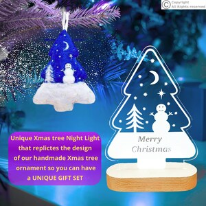 May include: A blue felt Christmas tree ornament with a white snowman, moon, and stars, paired with a clear acrylic night light in the same tree shape featuring a snowman, moon, stars, and the text Merry Christmas on a wooden base.