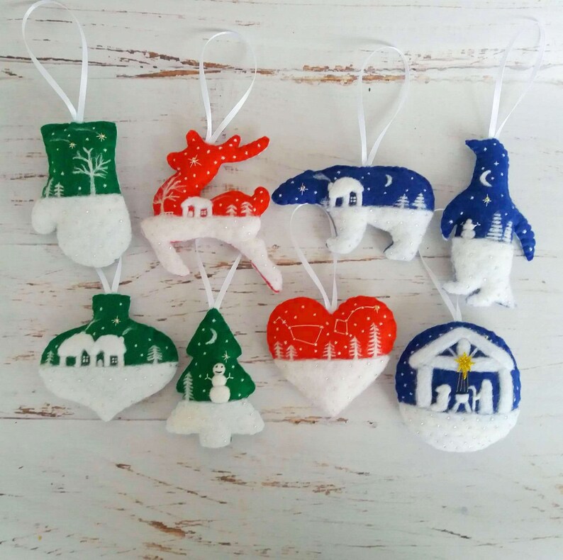 May include: A collection of eight felt Christmas ornaments featuring various shapes including a mitten, reindeer, polar bear, penguin, bauble, tree, heart, and nativity scene, each with white ribbon loops for hanging and detailed winter-themed embroidery.