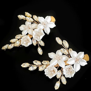 May include: Two gold hair clips, each decorated with white floral details and clear, sparkling crystals. The clips feature delicate, stylized flowers and leaf-shaped crystal embellishments, set against a black background.