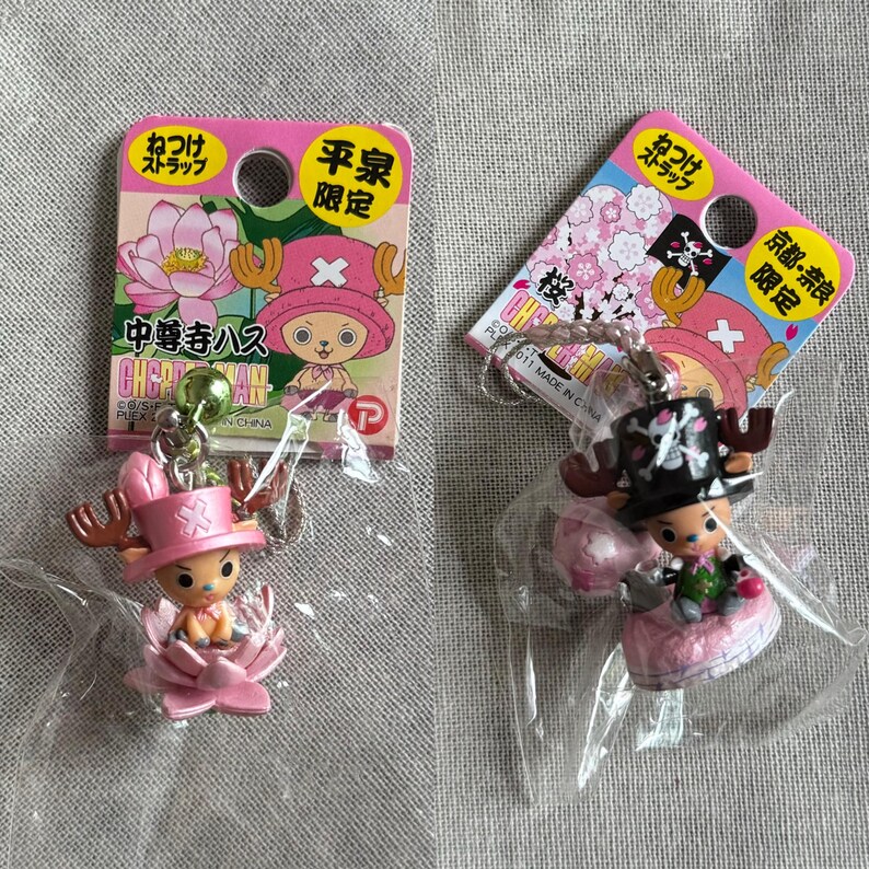 May include: Two One Piece anime character keychains. One features a pink hat and is seated in a pink flower. The other has a black hat with a skull design. Both have tags with Japanese text and illustrations.