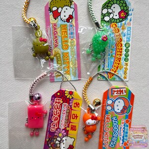 May include: Four Hello Kitty character charms on display cards. One is a green kiwi, one is a green sea grape cluster, one is a pink rectangular block, and one is an orange fruit. Each charm features a braided strap and a small metal bell.