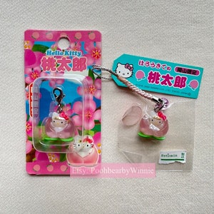 May include: A Hello Kitty charm featuring a pink peach with a Hello Kitty figurine inside. The charm is attached to a pink and white string with a pink Hello Kitty charm at the top. The charm is packaged on a pink and white card with the text "Hello Kitty 桃太郎" in Japanese.
