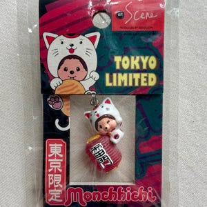 Op de afbeelding: Een Tokyo Limited Monchhichi sleutelhanger. De sleutelhanger heeft een kleine pop met een kattenhoed en een rood vat. De verpakking bevat de tekst "TOKYO LIMITED" en Japanse tekst.