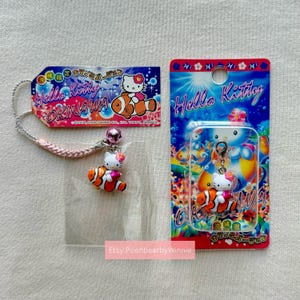 May include: Hello Kitty Okinawa charm set. The set includes a charm with a pink bow, a clownfish, and a tag with the text "Hello Kitty Okinawa". The charm is packaged in a clear plastic bag and a red and blue card.