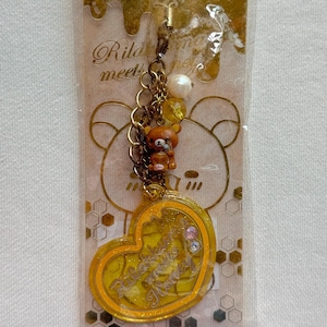 Può includere: Un ciondolo a forma di cuore giallo con la scritta "Rilakkuma meets Honey" e una piccola figura di orso marrone. Il ciondolo è attaccato a una catena d'oro con una perla e perline gialle. Lo sfondo presenta un'illustrazione di orso.