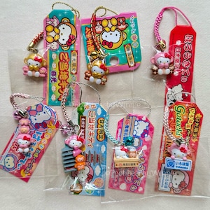 May include: A collection of Hello Kitty charms with tags. The charms feature Hello Kitty figures with various designs and accessories. The tags have Japanese text and colorful graphics. The charms are attached to a silver-colored string.