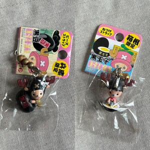 May include: Two One Piece anime character keychains. Each keychain features a small figure of Tony Tony Chopper with a black base. The keychains are attached to pink and white cardboard packaging with Japanese text.