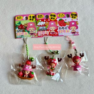 May include: Three One Piece character charms with red and white strawberry designs. Each charm is attached to a green and white string with a small bell. The charms are packaged on a pink and white card with Japanese text.