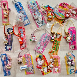 Puede incluir: Una colección de amuletos y etiquetas de Hello Kitty en varios diseños y colores. Los amuletos presentan a Hello Kitty con diferentes atuendos y poses, adjuntos a etiquetas coloridas con texto e ilustraciones japonesas. Algunas etiquetas tienen campanas.