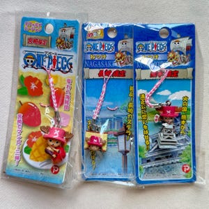 May include: Three One Piece character keychains in plastic packaging. The keychains feature the character Chopper in different poses, with a pink and white lanyard. The packaging includes Japanese text and illustrations.