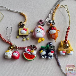 Può includere: Una collezione di ciondoli per telefono Hello Kitty con vari design, tra cui un quadrato giallo, un costume da pinguino rosa, un alimento marrone, due bambole daruma rosse e bianche, una fetta di anguria, due figure nere e scarpe dorate.