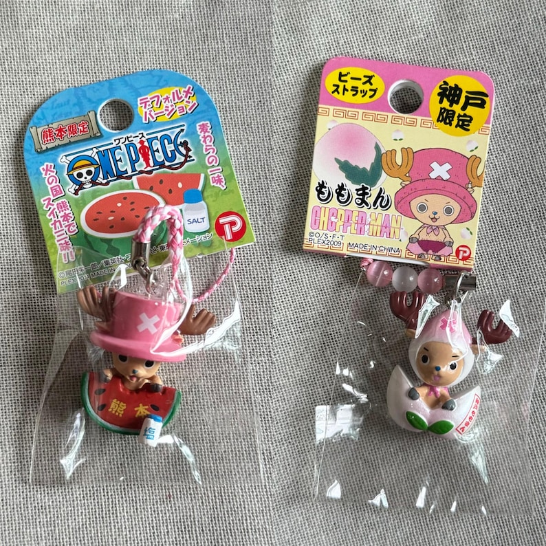 May include: Two One Piece Chopper Man character keychains. One is a figure of Chopper holding a watermelon slice, the other is Chopper in an egg. Both are in plastic packaging with Japanese text and pink and blue accents.