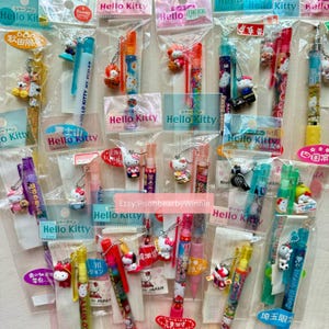 May include: A selection of Hello Kitty pens and mechanical pencils in clear packaging. The pens, in shades of red, blue, yellow, and purple, feature Hello Kitty charms and Japanese text. These stationery items are designed with a range of colours.
