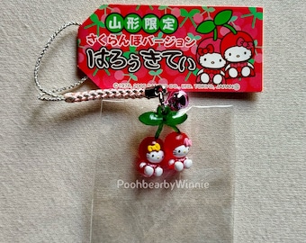 May include: A red tag with Japanese text and cherry illustrations is attached to a clear plastic card. Below, two Hello Kitty figures are designed as cherries. A silver cord and pink bell complete the charm.