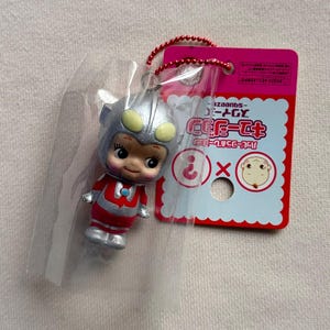 May include: A small, colorful figurine of a character in a clear plastic bag. The figurine is red, silver, and yellow, with a smiling face. A pink tag with Japanese text is attached to the bag with a red beaded string.