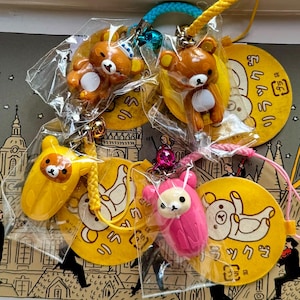 May include: A collection of Rilakkuma character keychains in various colors, including brown, yellow, and pink. Each keychain is attached to a yellow tag with Japanese text and a cartoon bear illustration. The keychains are packaged in clear plastic.