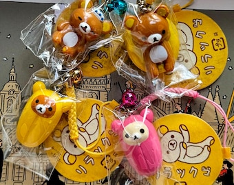 Può includere: Una collezione di portachiavi con personaggi di Rilakkuma in vari colori, tra cui marrone, giallo e rosa. Ogni portachiavi è attaccato a un'etichetta gialla con testo giapponese e un'illustrazione di orso dei cartoni animati. I portachiavi sono confezionati in plastica trasparente.