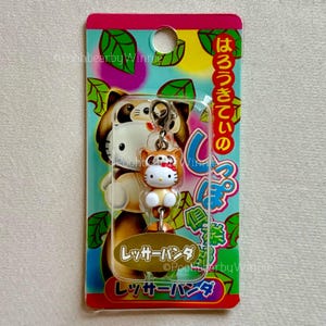 Puede incluir: Un llavero de Hello Kitty con una figura marrón y blanca con un lazo rojo, disfrazada de panda rojo. El llavero está sujeto a una tarjeta colorida con texto japonés y una ilustración de panda rojo.