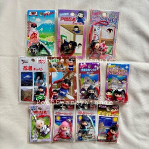 Può includere: Una collezione di 10 portachiavi colorati con piccole figurine di ninja e altri personaggi. Ogni portachiavi ha un laccio rosso e una piccola carta con testo e immagini giapponesi. Le carte presentano scene di monumenti giapponesi e architettura tradizionale.