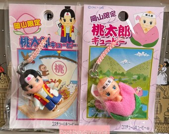 May include: Two Japanese keychain charms featuring the character Momotaro. One shows Momotaro in a boat, the other shows him emerging from a peach. Both are on pink and green cards with Japanese text.