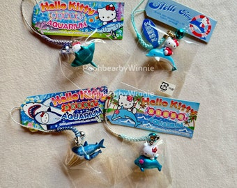 以下が含まれることがあります： ハローキティの水族館をテーマにしたチャームがいくつかあります。各チャームには、イルカやサメなどの海の生き物と一緒に小さなハローキティのフィギュアが描かれています。チャームには、ハローキティ水族館のロゴと日本語のテキストが付いたタグが付いています。