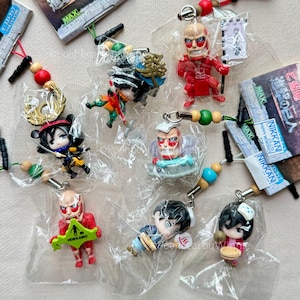 Puede incluir: Una colección de pequeños amuletos de personajes de anime coloridos, cada uno empaquetado individualmente. Los amuletos presentan varias figuras en poses dinámicas, con detalles como espadas, banderas y alimentos. Algunos amuletos tienen el texto "HOKKAIDO".