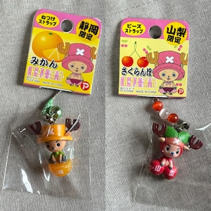 May include: Two Japanese anime character keychains. One keychain features a character with orange-colored antlers and a hat, sitting on an orange. The other keychain features a character with red antlers and a hat, sitting on a pair of cherries.