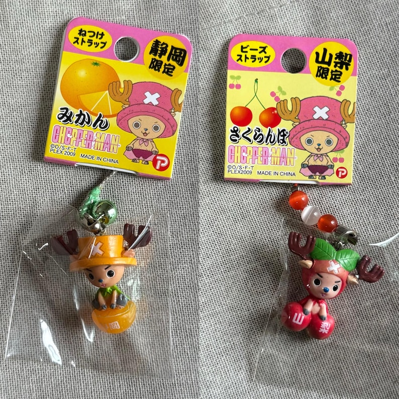 May include: Two Japanese anime character keychains. One keychain features a character with orange-colored antlers and a hat, sitting on an orange. The other keychain features a character with red antlers and a hat, sitting on a pair of cherries.