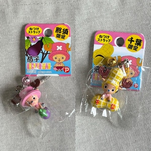 May include: Two collectible keychains featuring a cartoon character, each packaged on a pink card. One keychain depicts the character with an eggplant, the other with a peanut. The cards have Japanese text.