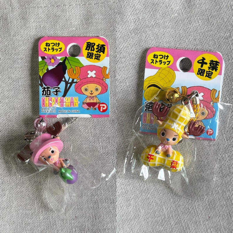 May include: Two collectible keychains featuring a cartoon character, each packaged on a pink card. One keychain depicts the character with an eggplant, the other with a peanut. The cards have Japanese text.