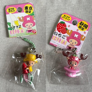 May include: Two Japanese anime character keychains. One keychain features a character with a banana hat and a watermelon, the other has a strawberry hat and is on a pink platform. Both keychains are in plastic packaging with pink tags.