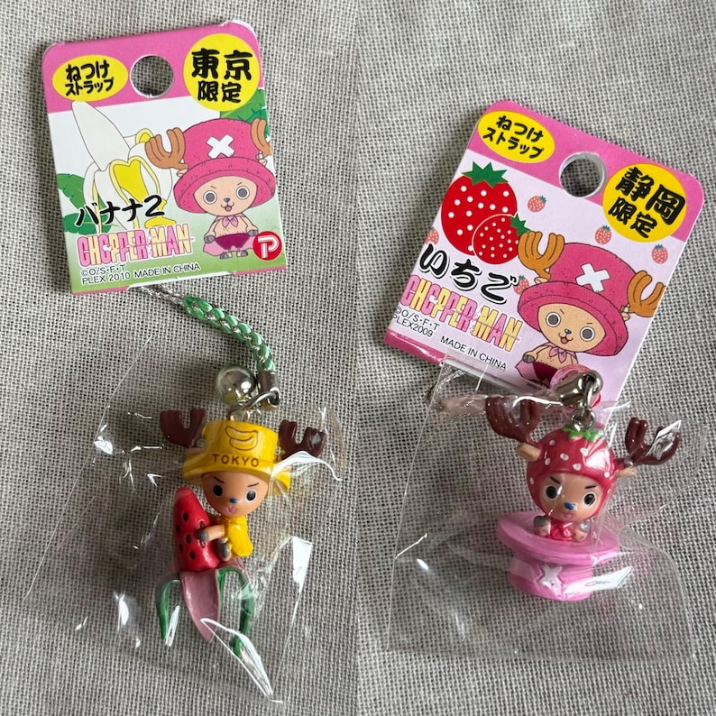 May include: Two Japanese anime character keychains. One keychain features a character with a banana hat and a watermelon, the other has a strawberry hat and is on a pink platform. Both keychains are in plastic packaging with pink tags.