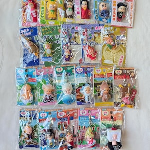 May include: A selection of small, packaged Japanese dolls, each with unique outfits and accessories. The dolls are in clear plastic packaging, with colourful backing cards featuring Japanese text and illustrations. The dolls vary in design, including traditional clothing and themed costumes.