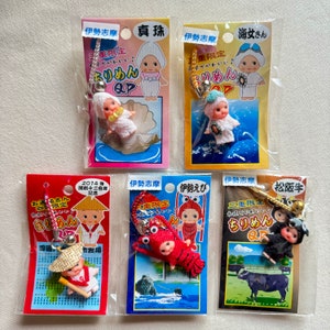 May include: A selection of Japanese souvenir keyrings featuring small doll figures. Each keyring has a different design, including a pearl, a diver, a lobster, and a cow. The keyrings are packaged in clear plastic with colourful backing cards.