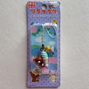 May include: A Rilakkuma character charm featuring a brown bear holding a pastel green and purple soft serve ice cream cone with a small yellow chick. The charm is attached to a string of translucent beads and packaged on a purple card.