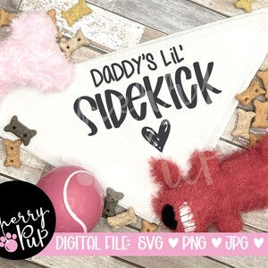 May include: White bandana with the text "Daddy's Lil' Sidekick" in black lettering with a small black heart. The bandana is surrounded by pink and red fuzzy toys and dog treats.