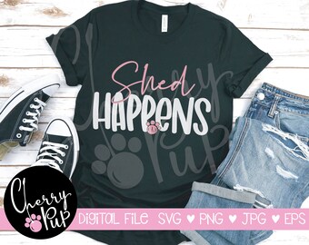 May include: A black t-shirt with the text "Shed Happens" in white and pink lettering. The text is surrounded by paw prints. The t-shirt is laid out on a wooden surface with a pair of black sneakers and a pair of blue jeans.