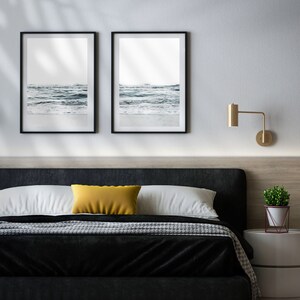 May include: A black bed frame with a black and white patterned blanket and a yellow pillow. Two framed black and white photos of ocean waves hang on the wall above the bed. A gold wall sconce is on the wall to the right of the bed. A small potted plant sits on a white nightstand.