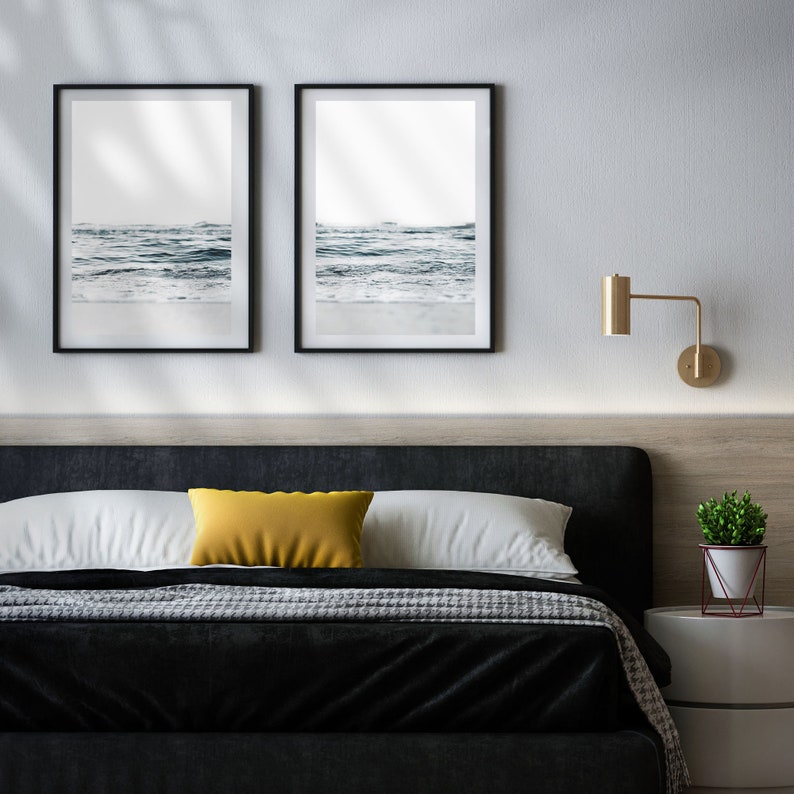May include: A black bed frame with a black and white patterned blanket and a yellow pillow. Two framed black and white photos of ocean waves hang on the wall above the bed. A gold wall sconce is on the wall to the right of the bed. A small potted plant sits on a white nightstand.
