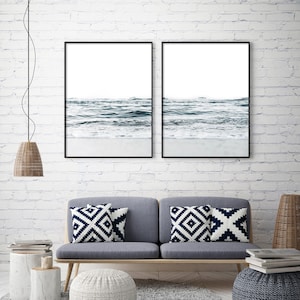 May include: Two framed black and white photographs of ocean waves crashing on a sandy beach. The photos are hung on a white brick wall above a gray couch with white and blue patterned throw pillows.
